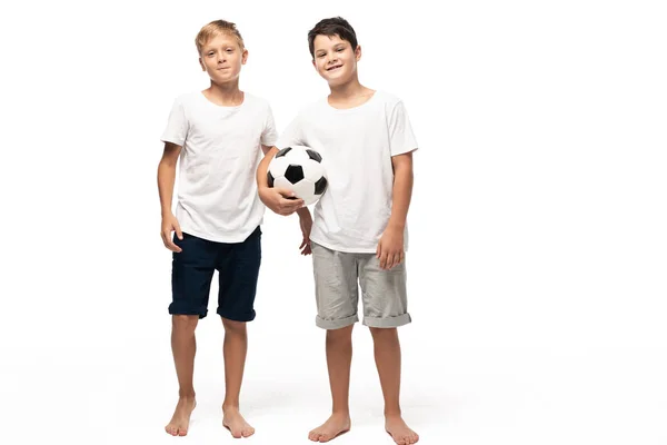 Cheerful boy holding soccer ball while standing near smiling brother on white background — Stock Photo