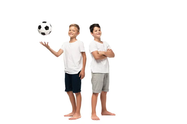 Веселый мальчик, играющий в футбол рядом с улыбающимся братом, стоящим со скрещенными руками на белом фоне — стоковое фото