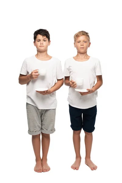 Dos hermanos serios mirando a la cámara mientras sostienen tazas de café sobre fondo blanco - foto de stock