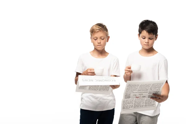 Zwei ernsthafte Jungen, die Zeitungen lesen, während sie Kaffeetassen isoliert auf weiß halten — Stockfoto