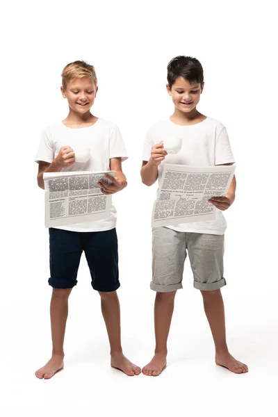 Dos hermanos sonrientes leyendo periódicos mientras sostienen tazas de café sobre fondo blanco - foto de stock