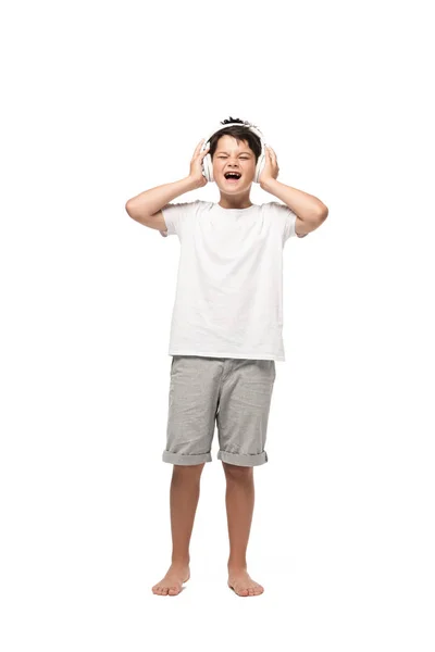Menino animado de mãos dadas em fones de ouvido e cantando enquanto ouve música no fundo branco — Fotografia de Stock