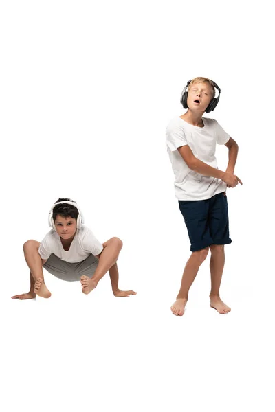 Excited boy singing in headphones near brother sitting in frog pose on white background — Stock Photo