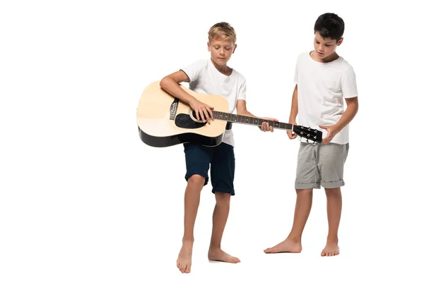 Menino de pé perto irmão tocando guitarra acústica no fundo branco — Fotografia de Stock