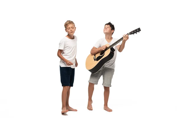 Chico de pie cerca de hermano tocando la guitarra acústica sobre fondo blanco - foto de stock