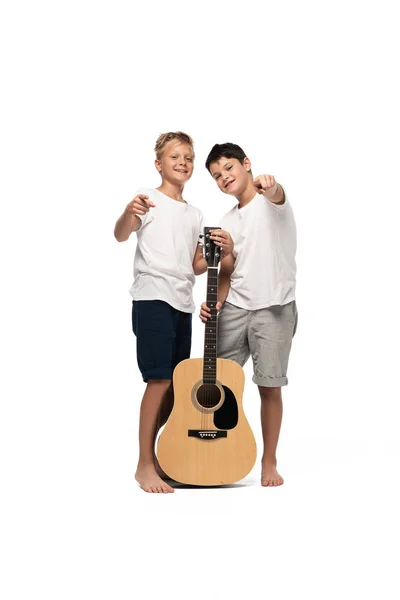 Dos hermanos alegres señalando con los dedos a la cámara mientras están de pie con la guitarra acústica sobre fondo blanco - foto de stock