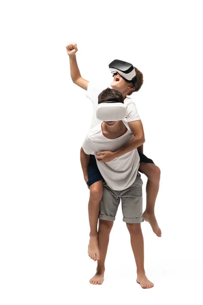 Allegro ragazzo piggybacking su fratelli indietro e mostrando sì gesto durante l'utilizzo di cuffie vr insieme su sfondo bianco — Foto stock