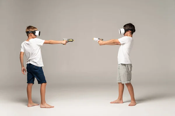 Visão de comprimento total de dois irmãos em fones de ouvido vr apontando um para o outro com armas de brinquedo em fundo cinza — Fotografia de Stock