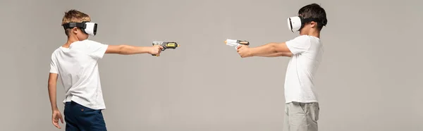 Панорамный снимок двух братьев в наушниках, нацеленных друг на друга игрушечными пушками, изолированными на сером — стоковое фото