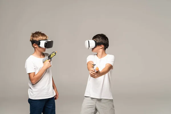 Два брата в гарнитурах VR смотрят друг на друга, держа игрушечные пистолеты изолированными на сером фоне — стоковое фото