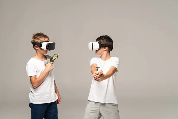 Zwei brüder in vr headsets schauen einander an, während sie spielzeugpistolen isoliert auf grau halten — Stockfoto