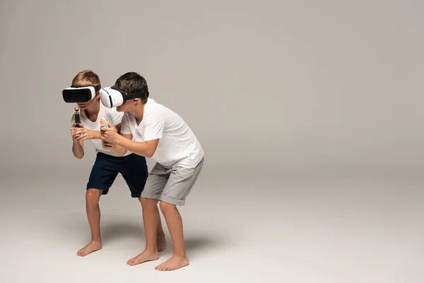 Two barefoot brothers in pajamas slinking with toy guns while using vr headsets on grey background — Stock Photo