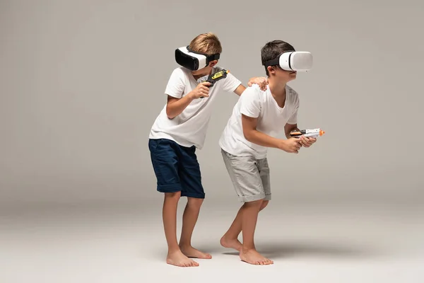 Deux frères pieds nus en pyjama tenant des pistolets jouets tout en utilisant des casques vr sur fond gris — Photo de stock