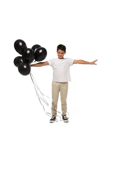 Chico serio de pie con las manos extendidas mientras sostiene globos negros sobre fondo blanco - foto de stock