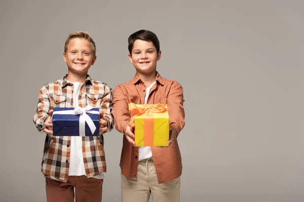 Dos hermanos felices sosteniendo cajas de regalo azules y amarillas aisladas en gris - foto de stock