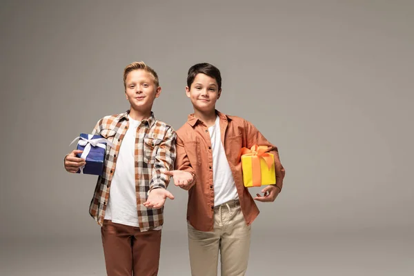 Два улыбающихся брата держат синие и желтые подарочные коробки, стоя с протянутыми руками, изолированными на сером — стоковое фото