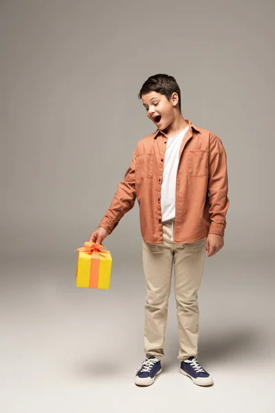 Menino animado segurando caixa de presente amarelo enquanto estava em fundo cinza — Fotografia de Stock