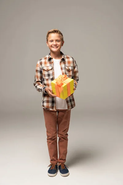 Niño alegre sosteniendo caja de regalo amarilla y sonriendo a la cámara sobre fondo gris - foto de stock