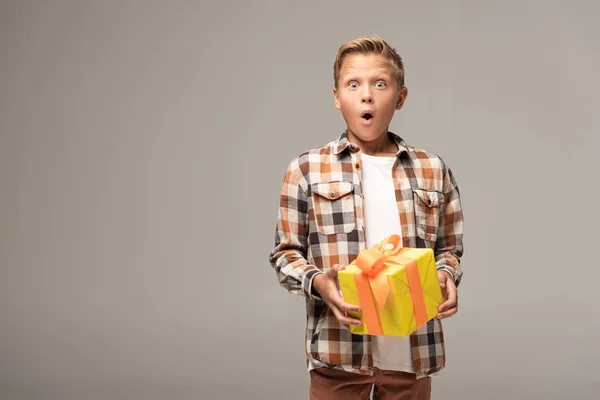 Shocked boy holding yellow gift box and looking at camera isolated on grey — Stock Photo