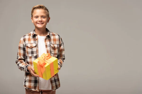 Niño feliz sosteniendo caja de regalo amarillo y sonriendo a la cámara aislada en gris - foto de stock