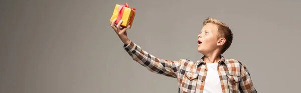 Panoramic shot of amazed boy holding gift box in outstretched hand isolated on grey — Stock Photo
