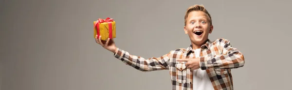 Панорамний знімок здивованого хлопчика, який вказує пальцем на подарункову коробку і дивиться на камеру ізольовано на сірому — стокове фото