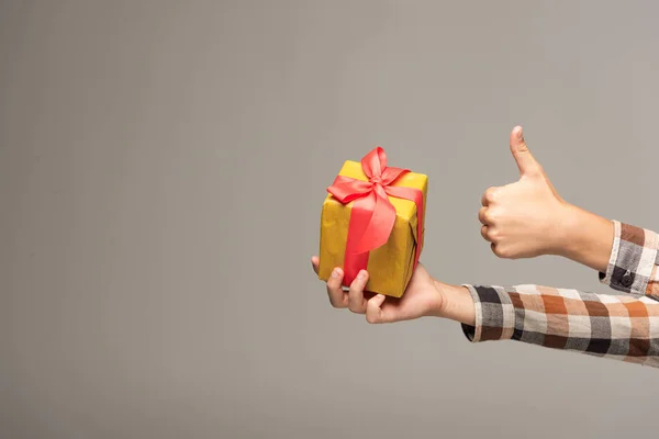 Cropped view of boy holding yellow gift box and showing thumb up isolated on grey — Stock Photo