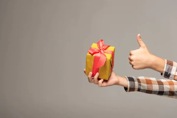 Vista ritagliata del ragazzo in possesso di scatola regalo gialla e mostrando pollice in alto isolato sul grigio — Foto stock