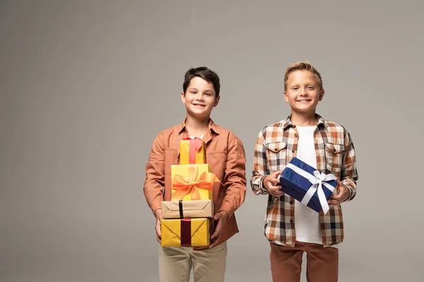 Dos hermanos felices sosteniendo cajas de regalo multicolores y sonriendo a la cámara aislada en gris - foto de stock