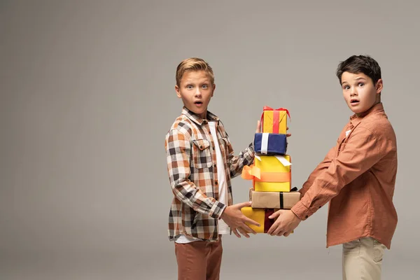 Two surprised brothers holding stack of multicolored gift boxes while looking at camera isolated on grey — Stock Photo