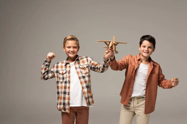 Dos hermanos alegres sosteniendo avión de juguete, mostrando sí gesto y mirando a la cámara aislada en gris - foto de stock