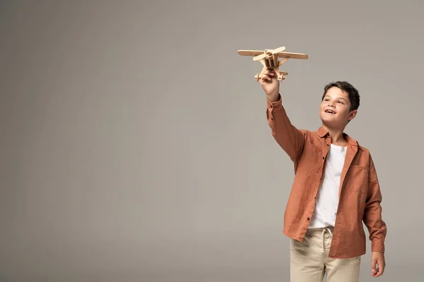 Happy boy holding wooden toy plane in raised hand isolated on grey — Stock Photo