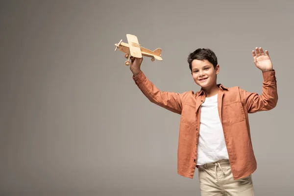 Smiling boy holding wooden toy plane and waving hand at camera isolated on grey — Stock Photo