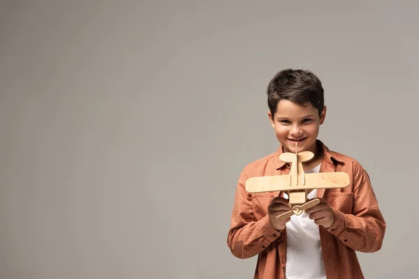 Cheerful boy showing wooden toy plane and smiling at camera isolated on grey — Stock Photo