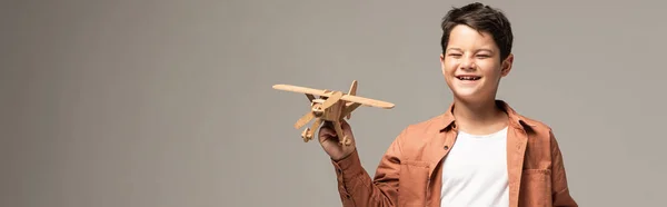 Панорамный снимок забавного мальчика, держащего деревянный игрушечный самолет, изолированный на сером — стоковое фото