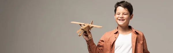 Plano panorámico de niño divertido sosteniendo avión de juguete de madera aislado en gris - foto de stock