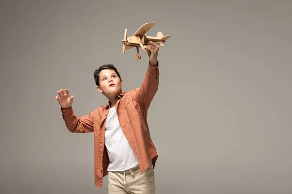 Amused boy holding wooden toy plane in raised hand isolated on grey — Stock Photo