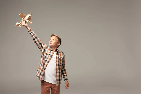 Glücklicher Junge mit Holzspielzeugflugzeug in erhobener Hand isoliert auf grau — Stockfoto