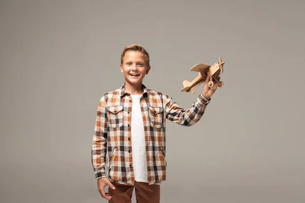 Glücklicher Junge hält Holzspielzeugflugzeug in der Hand und lächelt in die Kamera isoliert auf grau — Stockfoto