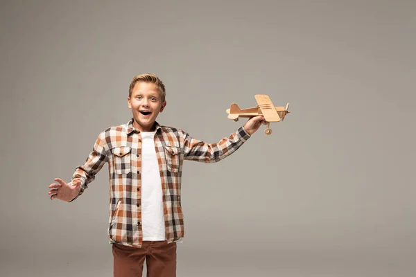 Niño excitado sosteniendo avión de juguete de madera y mirando a la cámara aislada en gris - foto de stock