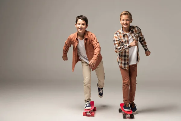 Dos chicos alegres montando tableros de peniques y sonriendo a la cámara sobre fondo gris - foto de stock