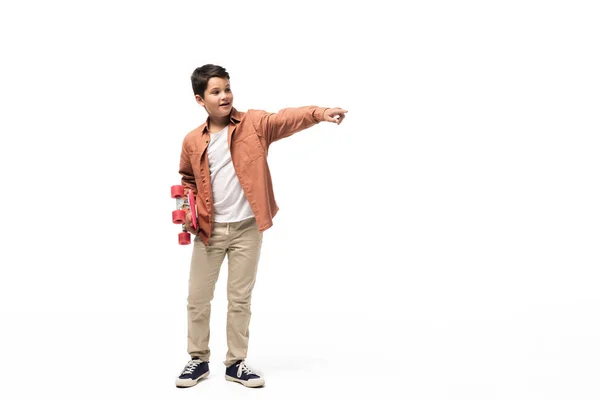 Sorrindo menino segurando penny board, olhando para longe e apontando com o dedo no fundo branco — Fotografia de Stock