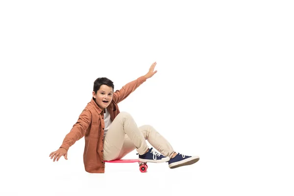 Alegre chico sentado en penny board y equilibrio con las manos extendidas sobre fondo blanco - foto de stock