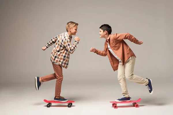 Side view of two cheerful brothers riding penny boards while looking at each other on grey background — Stock Photo