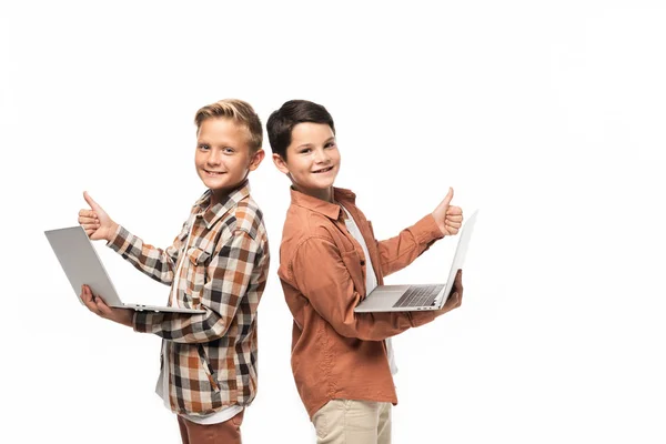 Dos hermanos sonrientes sosteniendo computadoras portátiles, mostrando los pulgares hacia arriba y mirando a la cámara aislada en blanco - foto de stock