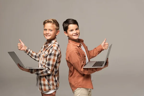 Two cheerful brothers holding laptops, showing thumbs up and looking at camera isolated on grey — Stock Photo