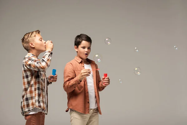 Мальчик раздувает мыльные пузыри рядом с изумленным братом, изолированным от серого — стоковое фото