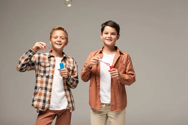 Two cheerful brothers holding bottles with soap bubbles and smiling at camera isolated on grey — Stock Photo