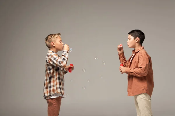 Vista lateral de dos hermanos soplando burbujas de jabón aisladas en gris - foto de stock