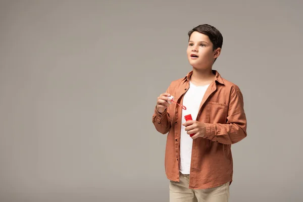 Verblüffter Junge hält Flasche mit Seifenblasen in der Hand und schaut isoliert auf grau — Stockfoto